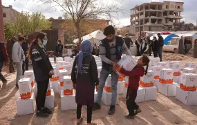 A young man and a young woman wear vests and stand among boxes of aid. The young man hands over a box of aid to girl. A boy stands in the middle of the frame, his back towards the camera.